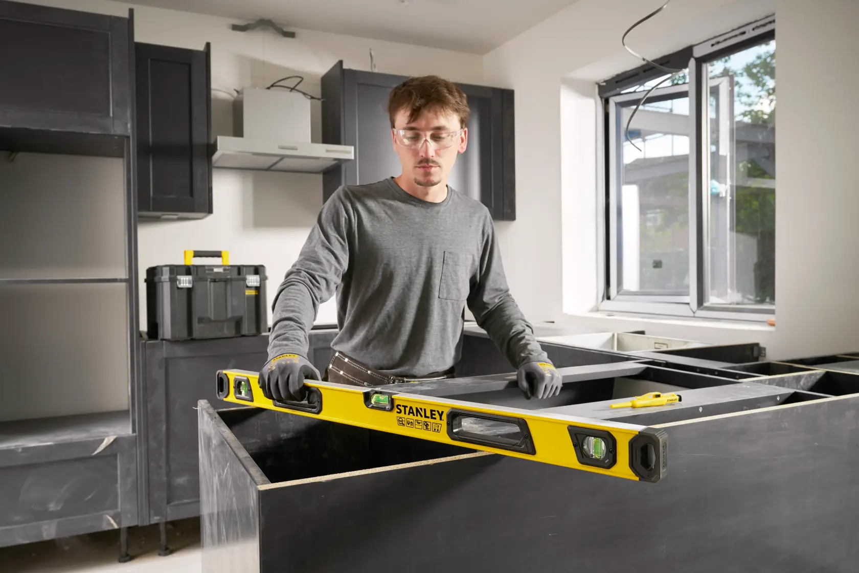 Person verwendet eine gelbe STANLEY Wasserwaage (Modell STHT41736-0) in einer im Bau befindlichen Küche. Die Person trägt Handschuhe und graue Kleidung und richtet eine Arbeitsplatte aus. Bauwerkzeuge und Materialien sind im Hintergrund sichtbar.