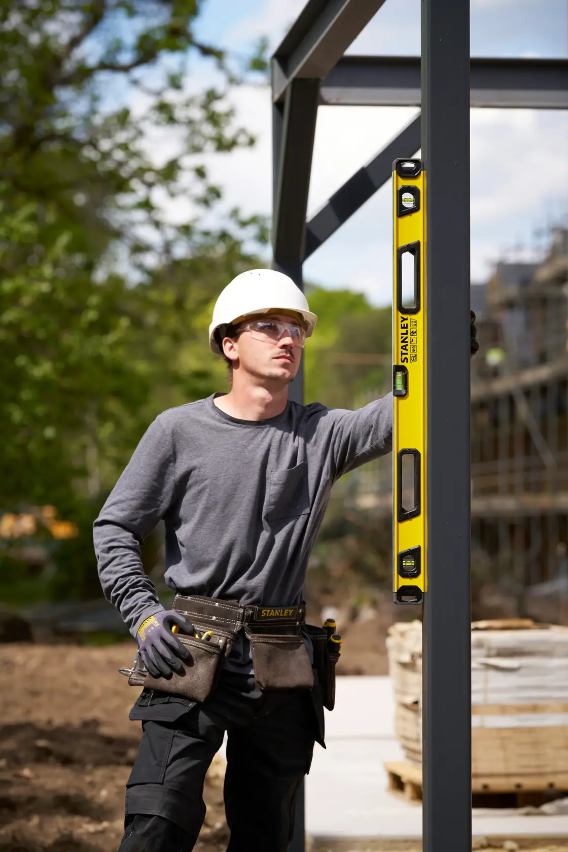 Ein Bauarbeiter verwendet eine gelbe STANLEY-Wasserwaage (SKU: STHT41736-0), um die Ausrichtung eines vertikalen Metallträgers auf einer Baustelle im Freien zu überprüfen. Der Arbeiter trägt einen Schutzhelm, ein graues Shirt, Handschuhe und einen Werkzeuggürtel. Im Hintergrund sind Bäume und Baumaterialien zu sehen.
