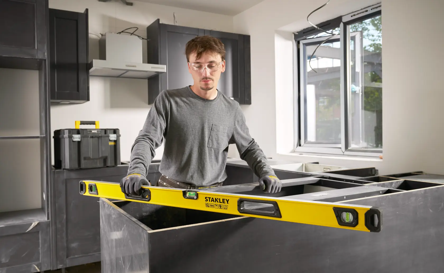 Eine Person verwendet eine Stanley-Wasserwaage, um eine Arbeitsplatte in einer im Bau befindlichen Küche zu vermessen und auszurichten; im Hintergrund sind Werkzeugkisten und Schränke sichtbar.