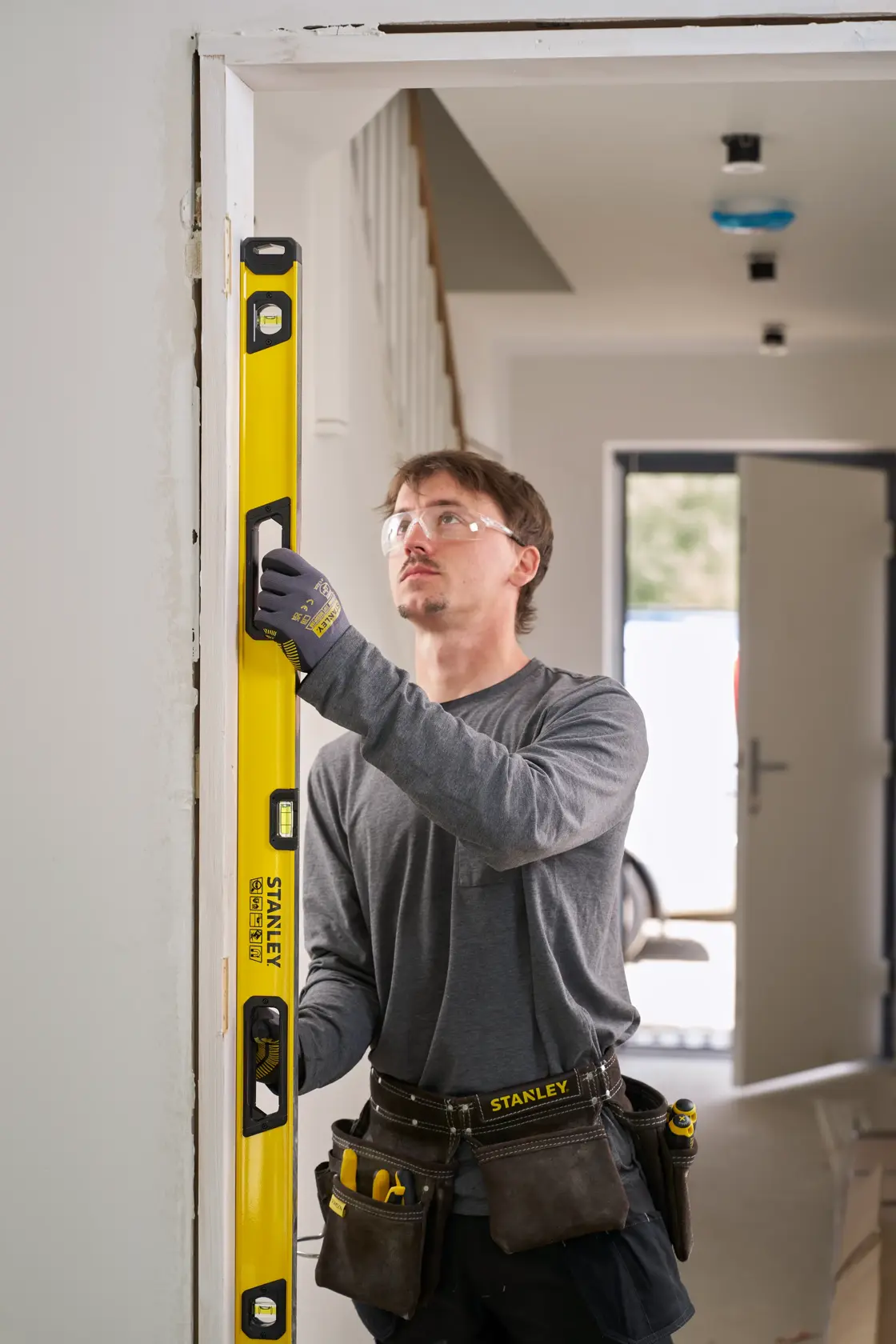 Eine Person mit Arbeitshandschuhen und Werkzeuggürtel prüft mit einer gelben STANLEY Wasserwaage die vertikale Ausrichtung eines Türrahmens in einem Innenbereich einer Baustelle. Das Gesicht der Person ist verschwommen.