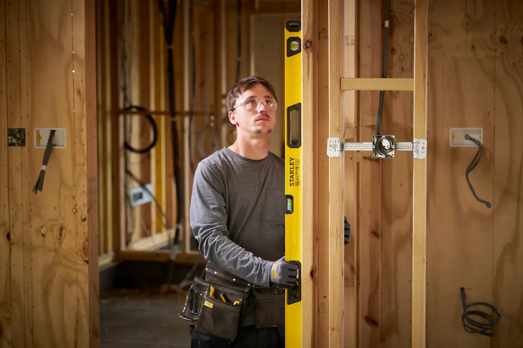 Eine Person mit Arbeitshandschuhen und Werkzeuggürtel verwendet eine gelbe STANLEY Wasserwaage, um die vertikale Ausrichtung eines Holzständerwerks auf einer Baustelle zu prüfen. Im Hintergrund sind unfertige Holzwände und Elektrokabel zu sehen.
