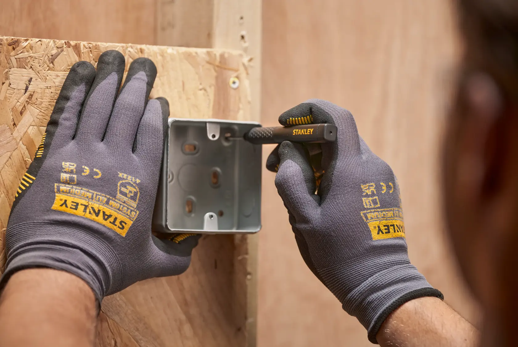 Hände mit STANLEY-Handschuhen markieren eine Metallbox auf Holz mit einem STANLEY-Stift.