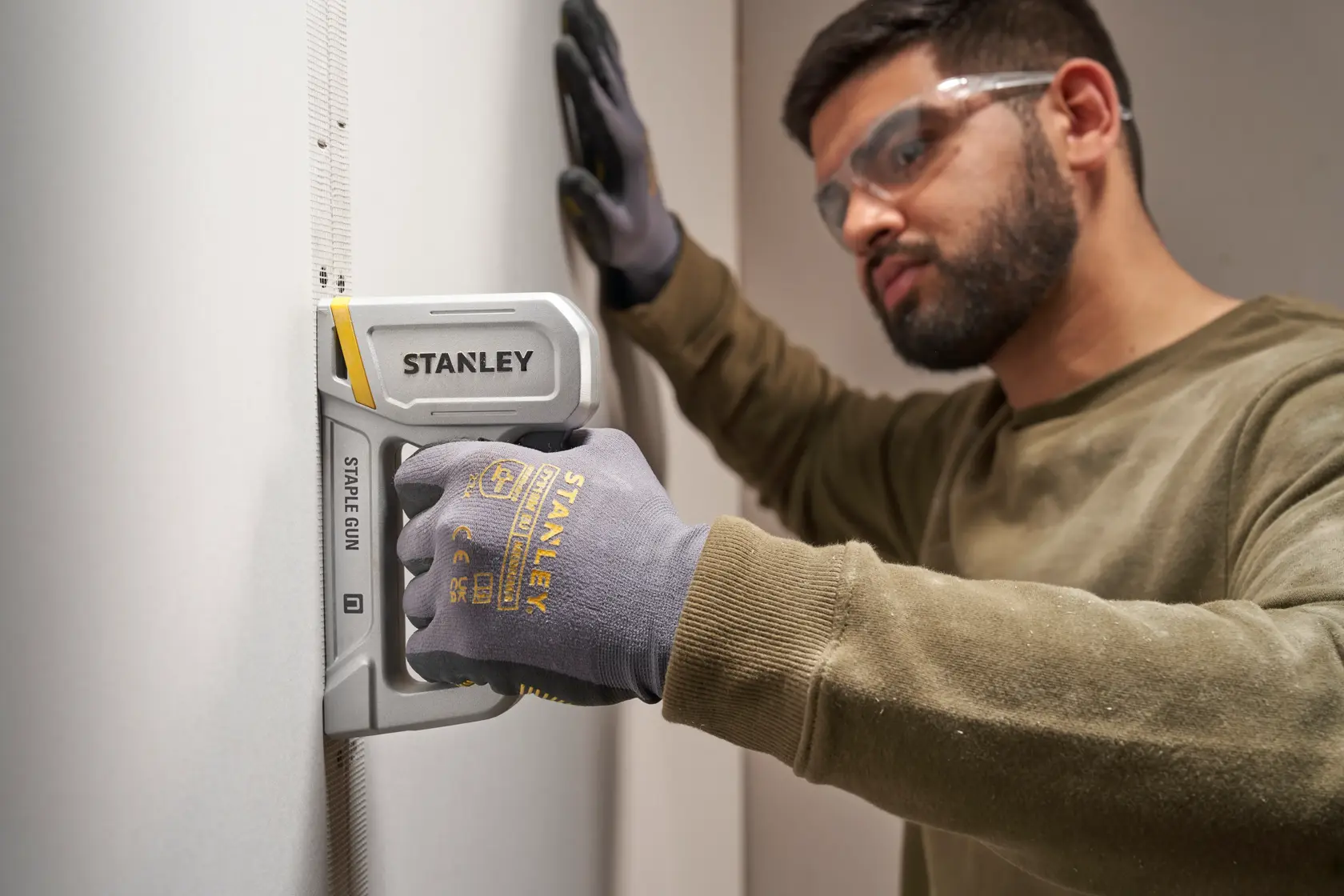 Foto der STANLEY STHT70150-0 Tackerpistole an der Wand, Person trägt STANLEY Handschuhe und hält das Werkzeug.