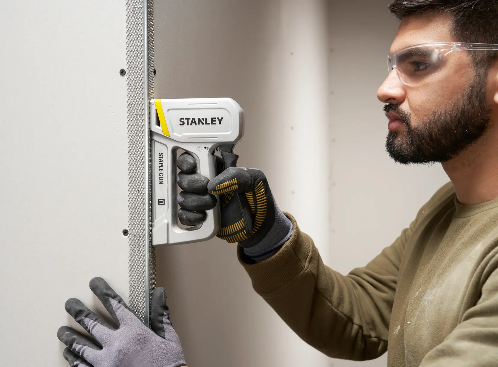 Fotografie des STANLEY STHT70150-0 Tackers, eingesetzt und von einer behandschuhten Hand an Trockenbau gehalten.