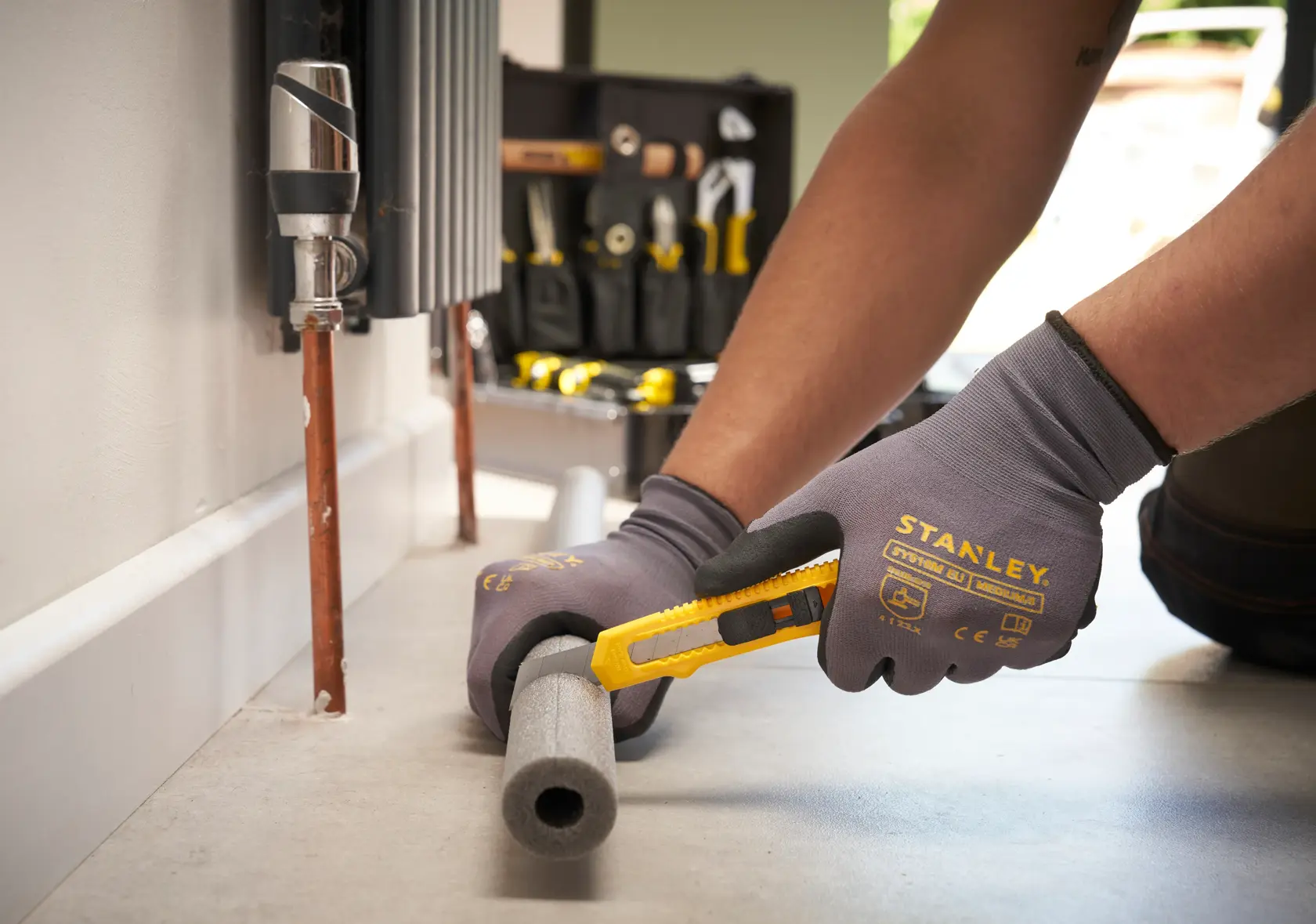 STANLEY STMT72986-1 Universalmesser schneidet Schaumrohrisolierung, Nahaufnahme, Handschuhe sichtbar, Werkzeugtasche im Hintergrund.