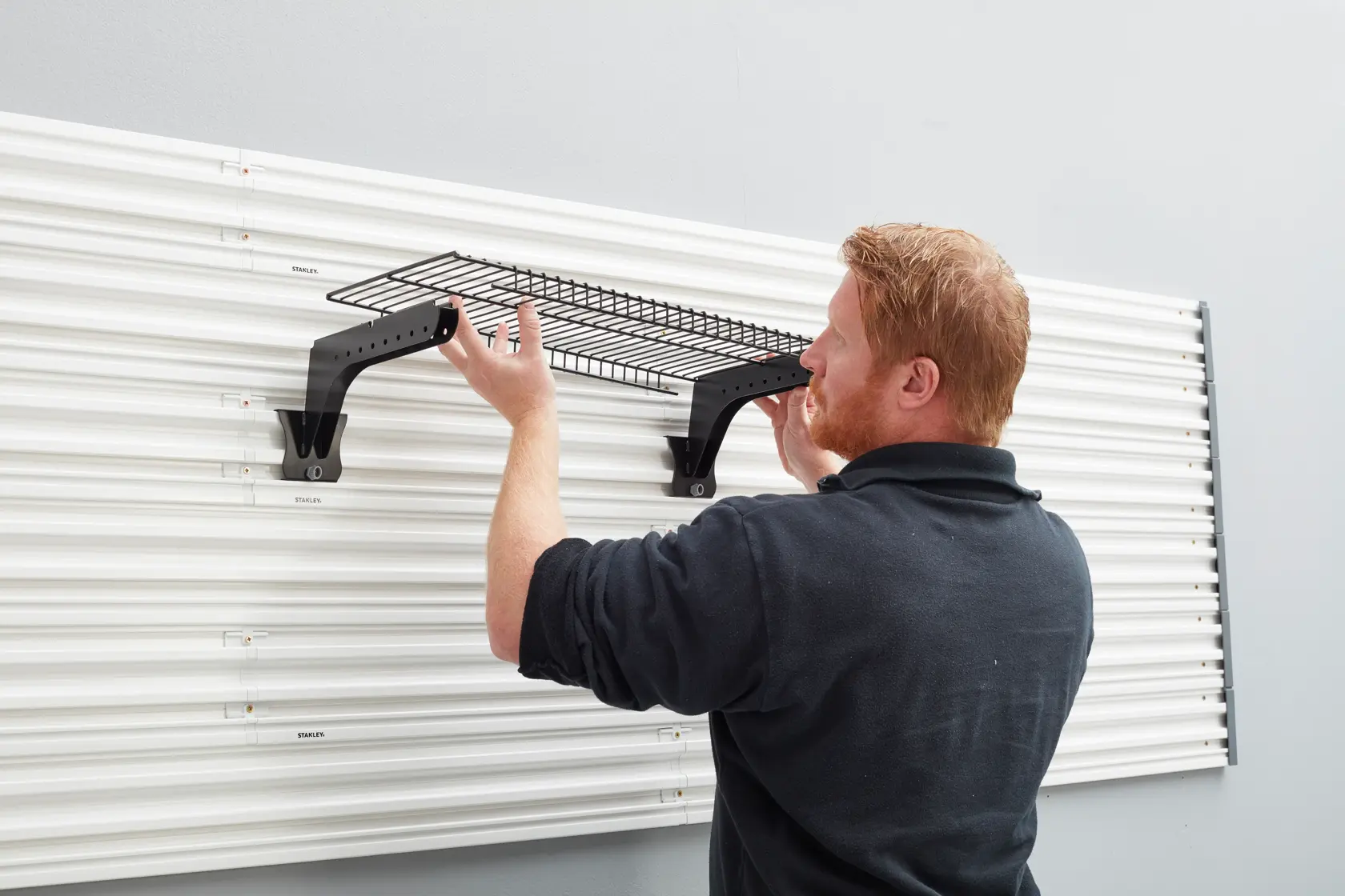 STANLEY STST82612-1 wall-mounted black wire shelf installation with brackets on white slatwall panel, side view.