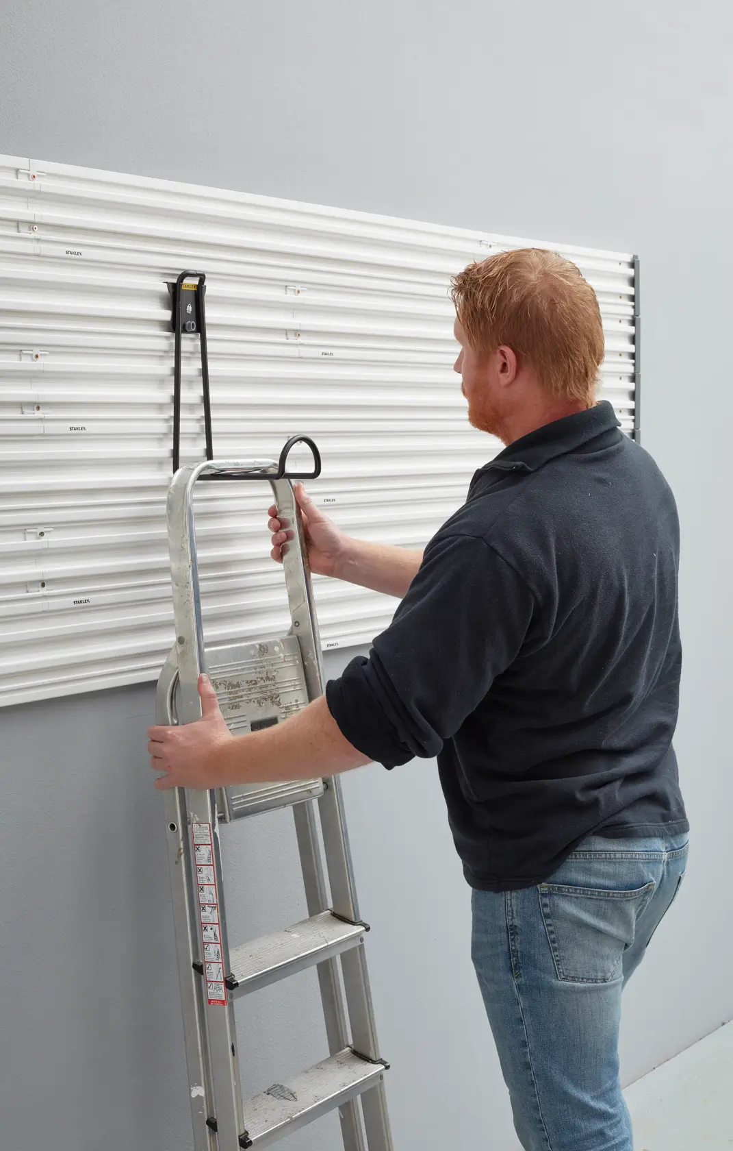 STANLEY STST82614-1 Wandhaken hält Metallleiter an weißer Lamellenwand, seitliche Aufnahme.