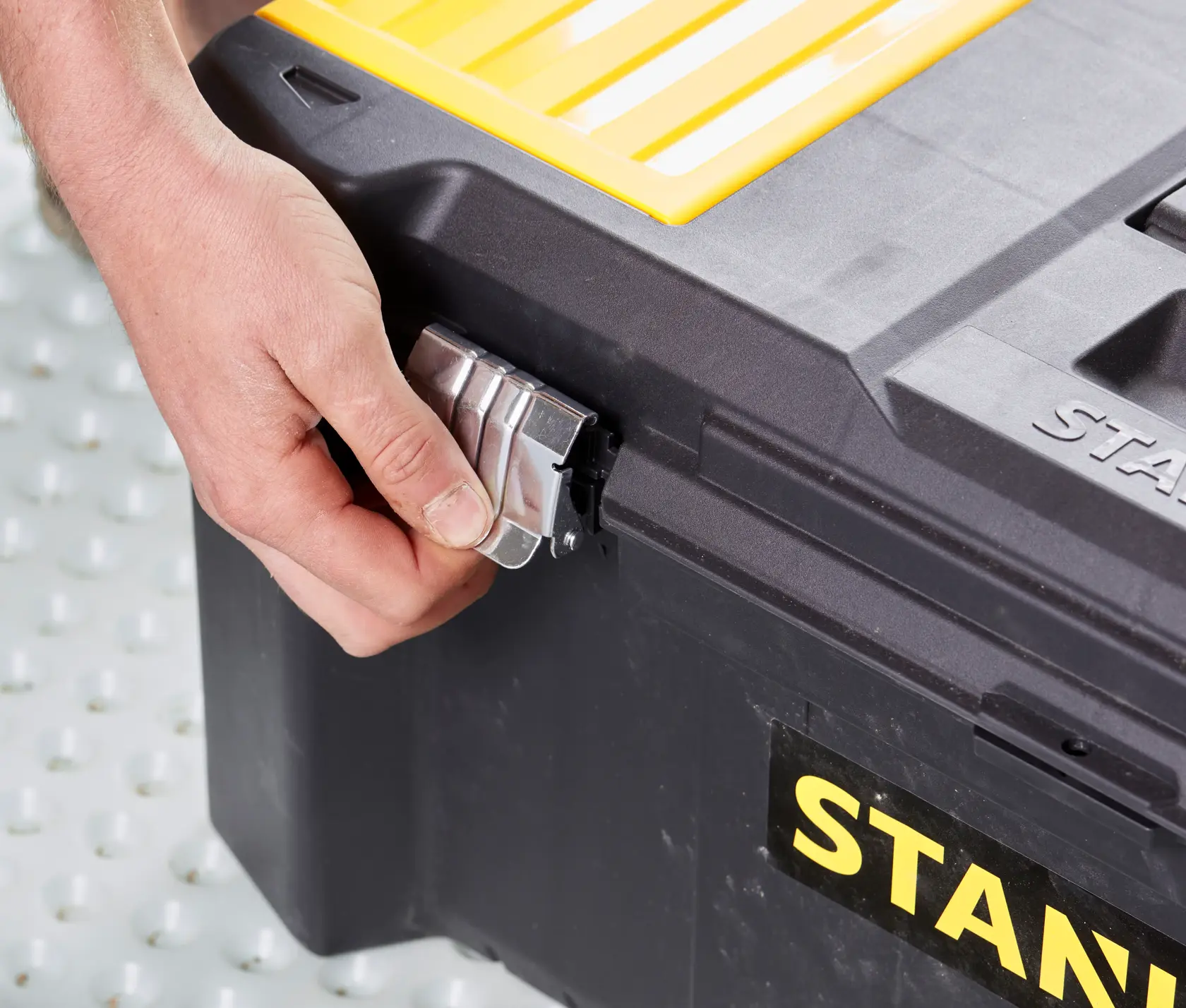 STANLEY STST82976-1 Werkzeugkasten Seitenansicht, Hand öffnet Metallschließe, schwarze Box, gelber Deckel.